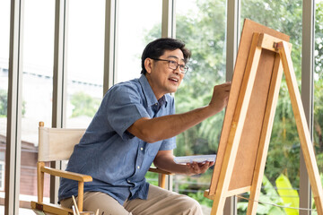 Indoor shot of senior Asian men using brush and oil color to paint on painting canvas. With smile on his face. Happy retirement concept for active senior. With Blurred living room background.