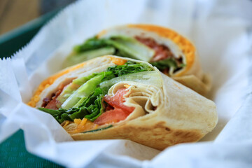 A close-up image of a healthy turkey wrap cut in half with cheese, tomatoes, and lettuce. Selective focus at the front half of the wrap.  
