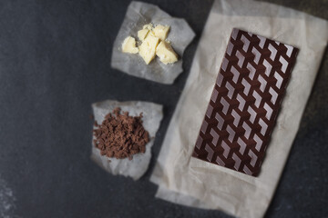 A bar of natural milk vegan chocolate with cocoa and cocoa butter ingredients on parchment paper on a black background. Handmade chocolate. Healthy dessert. Copy space.