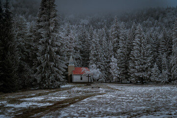 inter landscape with a small chapel surrounded by snow-covered fir trees in the morning fog