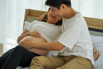 Asian pregnant woman and boyfriend looking at clothes for new born baby on bed at home