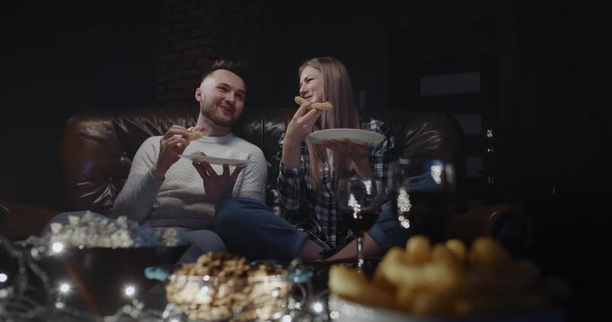 Happy Millennial Couple Sitting On Couch Having Fun Watching TV Show Or Movie At Home In Evening. Handsome Man And Attractive Woman Eating Pizza, Talking And Smiling Each Other. Wide Angle Dolly Shot