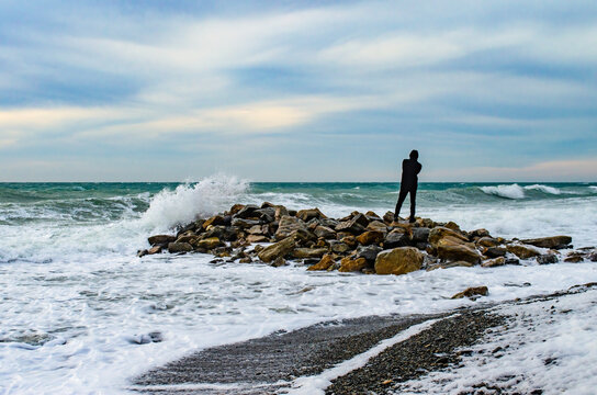 Splashing wave on the Black sea in the evening. A man stands on a stone pier with his back to the viewer and looks at the waves. A man takes pictures of the waves on his smartphone.