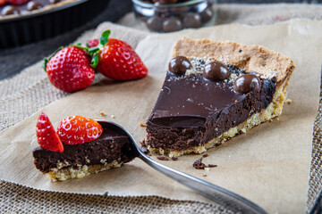Dessert spoon cutting chocolate tart or pie slice topped with strawberries, chocolate candy and almonds. Selective focus on cake, blurred background. Delicious gluten-free homemade crust or tartlet.