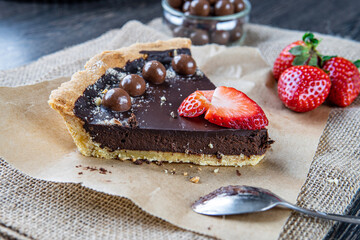 Chocolate tart or pie slice topped with strawberries, chocolate candy and almonds on rustic brown cloth. Dessert spoon in background. Delicious gluten-free homemade crust or tartlet. Side view.