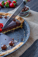 Kitchen knife slicing chocolate tart or pie topped with strawberries, chocolate candy and almonds. Delicious gluten-free homemade crust or tartlet. Side view.