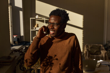 Technology, job and communication concept. Indoor image of attractive young African man freelancer discussing business issues on phone, calling partner, posing at home with laptop in background