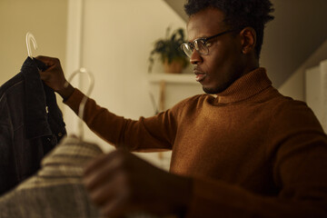 Clothing, shopping, wardrobe, style and fashion concept. Portrait of worried indecisive young dark skinned guy in sweater holding hangers, choosing shirt, getting dressed for date, feeling nervous