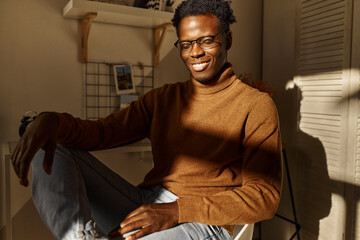 Horizontal image of joyful successful young African American businessman working from home in stylish sunlit room, smiling happily with his eyes closed, sitting comfortably in relaxed posture