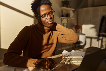 Tired young dark skinned male teacher in glasses massaging neck having exhausted look, working from home using laptop, giving online lessons. Sleepy black businessman sitting at desk with papers