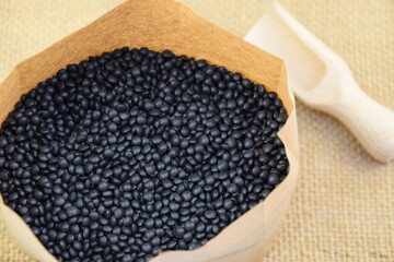 Black lentil, beluga lentil in paper bag on jute fabric background, wooden spoon blurred