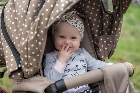 Happy smiling emotional eight-month-old blue-eyed girl sit in stroller on walk and wait for mom. Small and cute baby with headband on head sitting in pram. Concept of proper upbringing and childhood