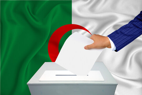 Elections In The Country - Voting At The Ballot Box. A Man's Hand Puts His Vote Into The Ballot Box. Flag Algeria On Background.