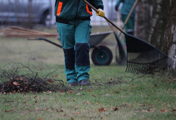 Eine nicht erkennbare G&auml;rtnerin in gr&uuml;ner Arbeitskleidung harkt &Auml;ste und Gras mit Laubrechen 