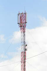 A red-white cell tower made of metal with wires against a blue sky with clouds