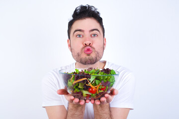 Charming cheerful Young handsome Caucasian man holding a salad bowl against white background making up plan in mind holding hands together, setting up an idea. Look askance