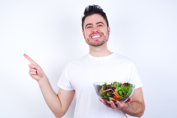 Young handsome Caucasian man holding a salad bowl against white background looking at camera indicating finger empty space sales