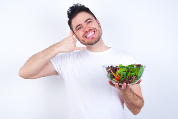 Young handsome Caucasian man holding a salad bowl against white background smiling doing phone gesture with hand and fingers like talking on the telephone. Communicating concepts.