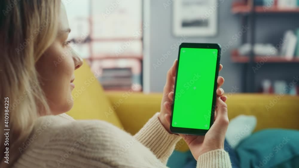 Aufkleber Beautiful Authentic Female Using a Smartphone with Green Screen Mock Up Display at Home Living Room while Lying on a Couch Sofa. She's Browsing the Internet and Checking Videos on Social Networks.