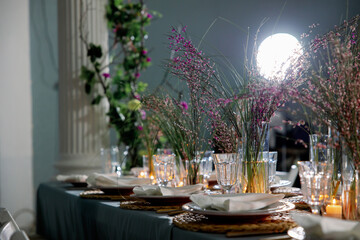 Table decorated with genista flowers and bergrass. Serving with wicker coasters for plates, napkins, burning candles and glasses. Decorations and table for wedding and dinner parties.