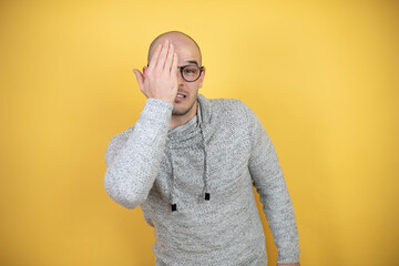 Young bald man wearing glasses over yellow background covering one eye with hand, confident smile...