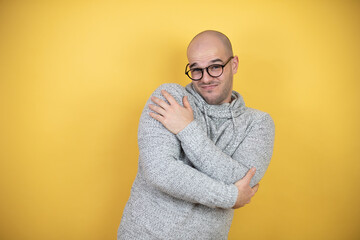 Young bald man wearing glasses over yellow background hugging oneself happy and positive, smiling...