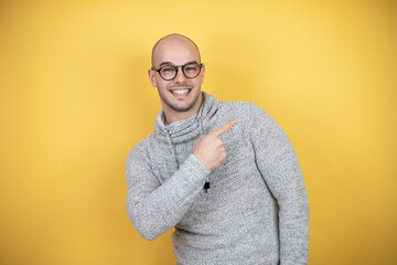 Young bald man wearing glasses over yellow background surprised and pointing with hand and finger...