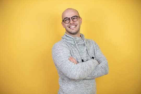Young bald man wearing glasses over yellow background with a happy face standing and smiling with a confident smile showing teeth with arms crossed