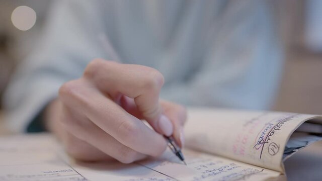 Close-up of slim female Caucasian hand writing in slow motion indoors. Unrecognizable young woman handwriting in workbook sitting at table. Student or businesswoman lifestyle.