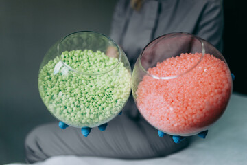 Master of epilation is holding two round glass vases with pink and green wax granules for waxing. The concept of depilation, epilation, waxing, shugaring.