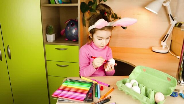A girl with rabbit ears on her head paints white eggs with felt-tip pens at the table. Preparation for Easter, educational activities for children, how to interest a child? Easter bunny, DIY craft