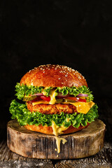 Delicious homemade grilled burger with turkey cutlet with cheese, cheese sauce, tomato, cucumber, salad on wooden board, on dark background. Close up, selective focus with copy space, vertical frame