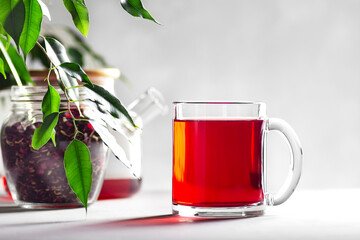 Transparent mug with red tea on a gray background, next to a jar with hibiscus and a teapot,...