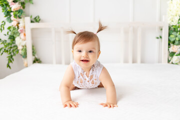 funny baby girl six months old sitting in a bright beautiful room on a white bed in a lace bodysuit looks away and smiles