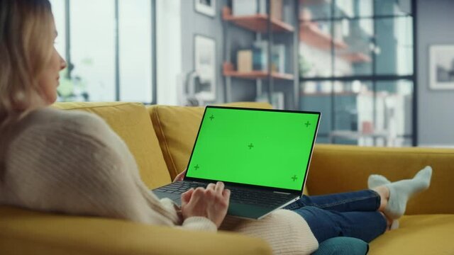 Beautiful Caucasian Specialist Working on Laptop with Green Screen Mock Up Display at Home Living Room while Lying on a Couch Sofa. Freelance Female Chatting Over the Internet on Social Networks.