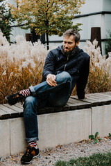 Handsome middle-aged Caucasian man portraited in the city, sitting on a bench with grey and black hair having a friendly look on his face, wearing a blue jacket and jeans. Fall colourful background.