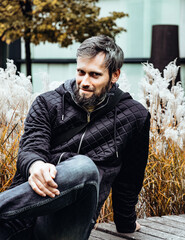Handsome middle-aged Caucasian man portraited in the city, sitting on a bench with grey and black hair having a friendly look on his face, wearing a blue jacket and jeans. Fall colourful background.