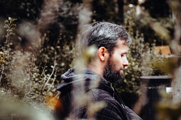 Handsome middle-aged Caucasian bearded man portrayed in the city, sitting on a park bench, holding a cell phone, with grey and black hair having a friendly look on his face, wearing city clothes.