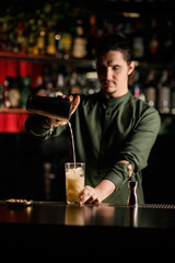 young bartender pours cocktail from steel shaker into crystal glass
