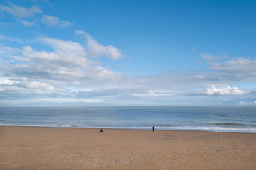 A winter beach on a blue sky day with fluffy white clouds. A few unrecognisable people are walking on the beach.