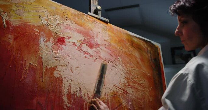 Cinematic shot of female creative artist painter concentrated and inspired painting colorful abstract picture with paint tool and oil colors on canvas while working on new masterpiece for exhibition.