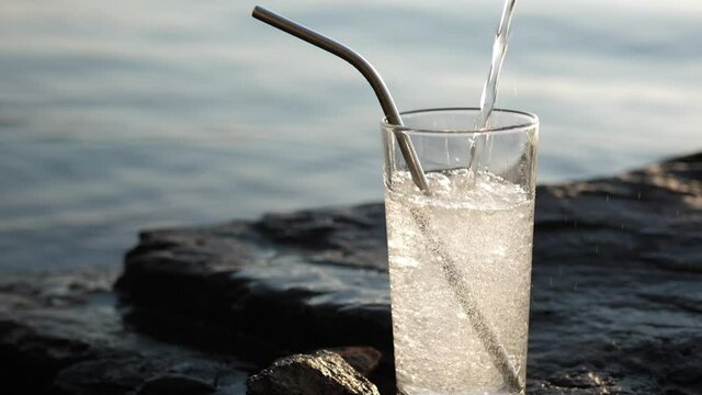 Soda water pours into a tall glass with a reusable metal cocktail tube on a rocky seashore slow motion. The soft drink sizzles, bubbles, and splashes.