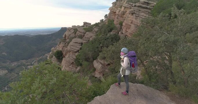 Mujer encima de monta&ntilde;a pedregosa con mochila y m&aacute;scara facial mirando infinito