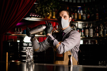 bartender in apron and mask and gloves stands behind the bar and holds steel shaker
