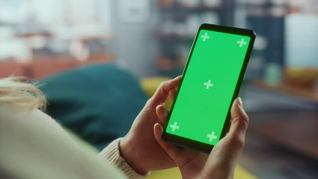 Close Up of an Authentic Female Using Smartphone with Green Screen Mock Up Display at Home Living Room while Lying on a Couch Sofa. She's Browsing the Internet and Checking Videos on Social Networks.
