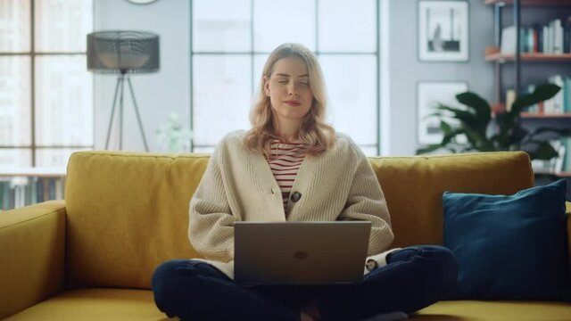 Beautiful Caucasian Specialist Working on Laptop Computer at Stylish Home Living Room while Sitting on a Cozy Couch Sofa. Freelance Female Browsing Internet, Using Social Networks, Having Fun in Flat.