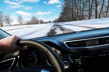 Car dashboard with driver's hand on the steering wheel with winter road in motion against a sky with clouds