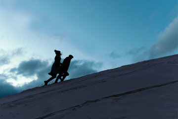 A young couple runs uphill against the evening blue sky with clouds. Black silhouettes of people....