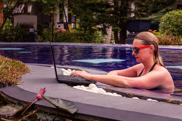 An attractive young female in orange sunglasses and wet long hair swims in the pool and works on a laptop. The girl looks at the computer screen. Freelance work on vacation.