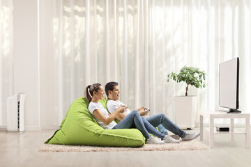 Man and woman sitting on green bean bag armchairs and playing video tv games at home
